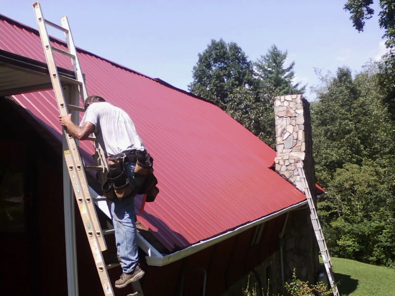 Roofer on a ladder beside a red metal roof and stone chimney — Soffit & Fascia Repair by Roofing Brothers in Greycliff
